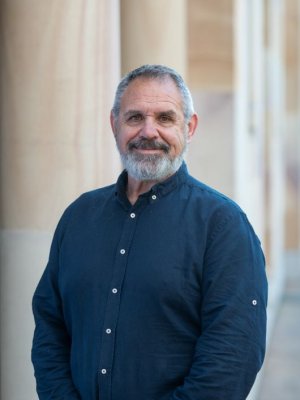 A man with greying hair and beard wearing a navy shirt stands with sandstone columns behind him.